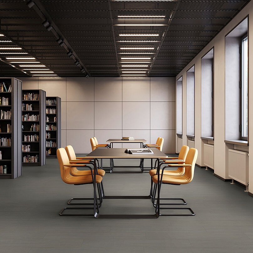 Modern reading area with clean lines – recycled elastic flooring in a dark grey, finely textured design for a calm, durable and easy-care surface. Library room with bookshelves, tables and yellow chairs on recycled elastic flooring with a modern dark grey texture.